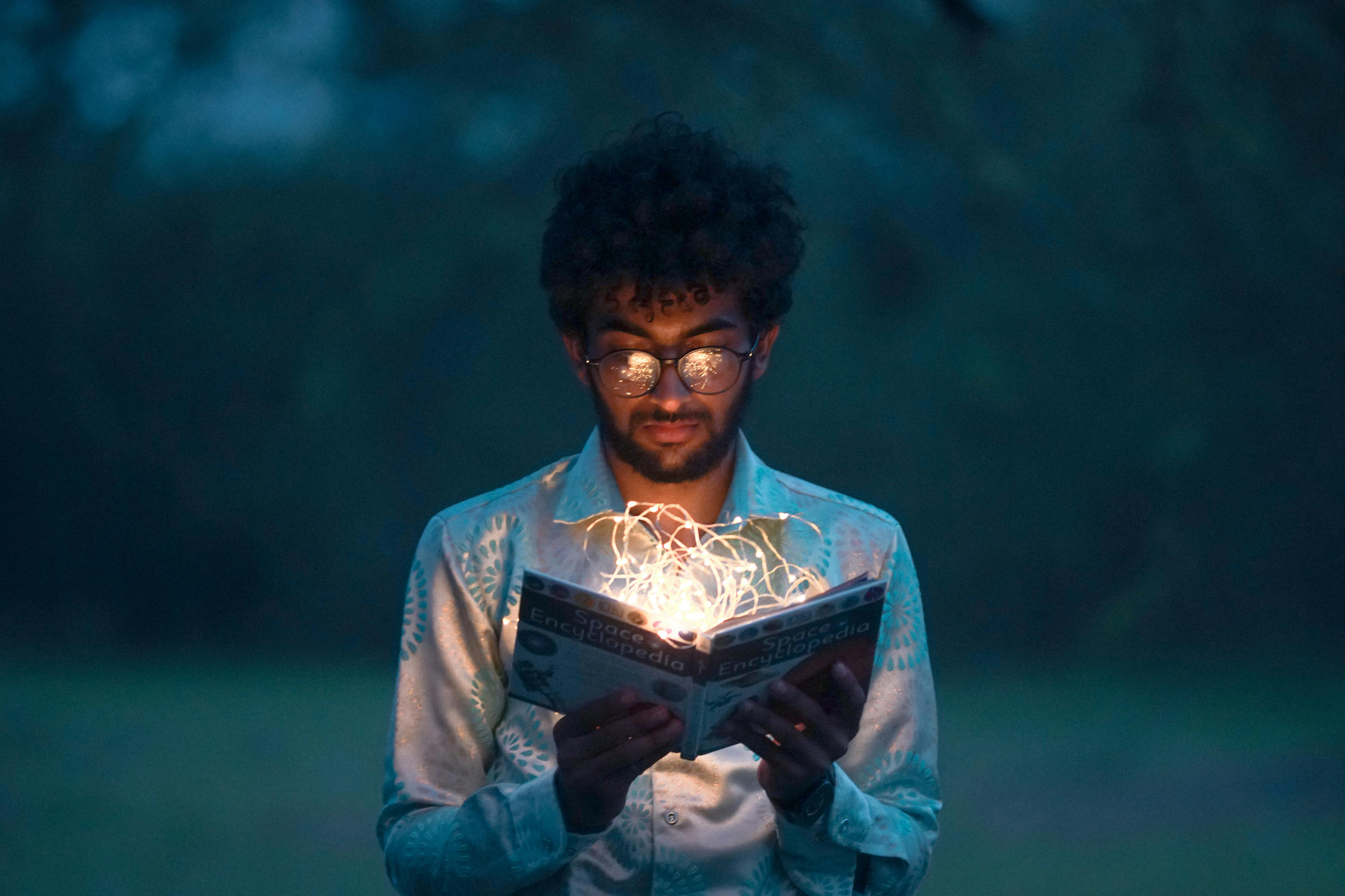 Hombre de tez oscura con gafas leyendo un libro del cual salen una especie de luces